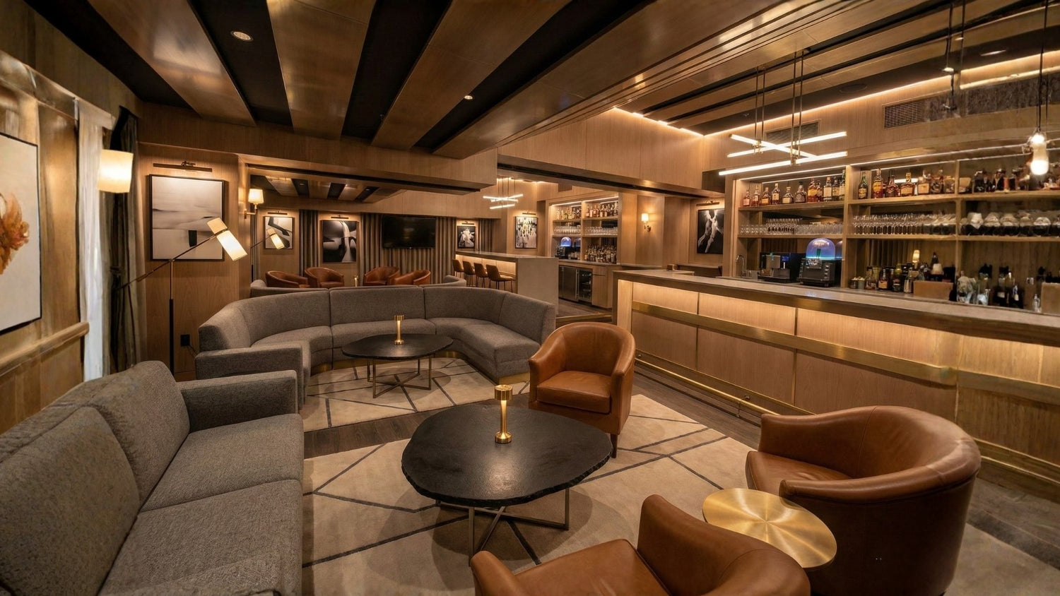 Salon bar moderne avec boiseries, canapé courbe gris, fauteuils en cuir cognac et bar intégré éclairé.
